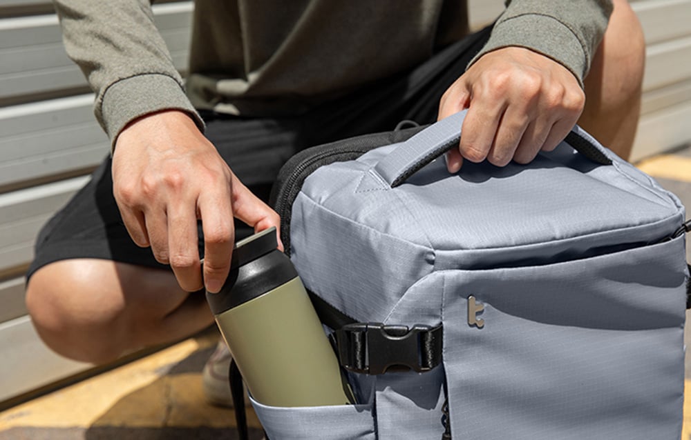 A person taking a water bottle from the side pocket of a gray backpack