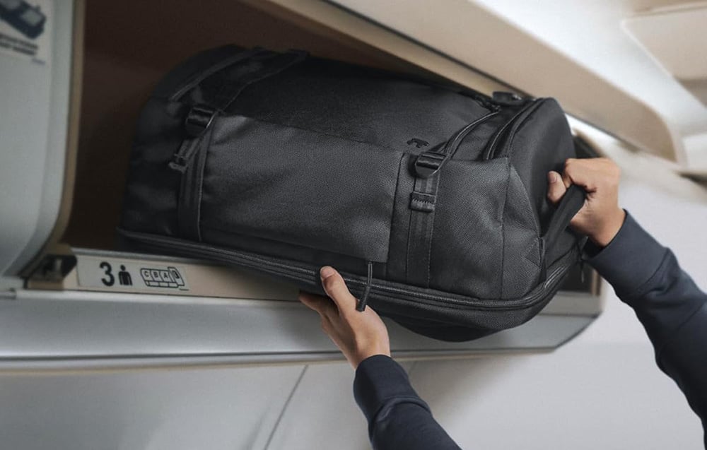 Ręce wkładające czarny plecak Tomtoc Navigator T67 do schowka bagażowego w samolocie