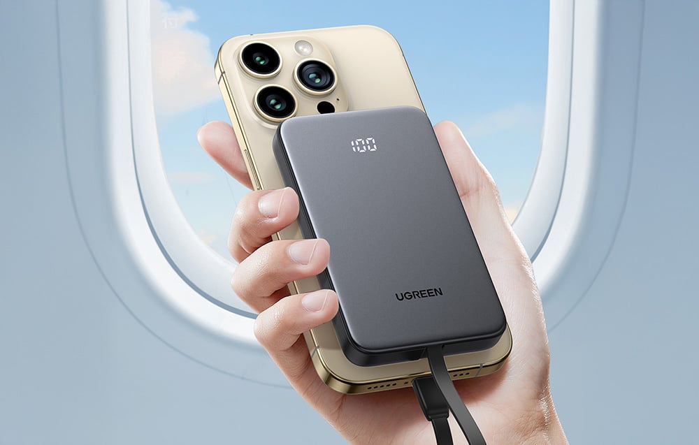 Dłoń trzymająca powerbank podłączony do smartfona na tle okna samolotu.