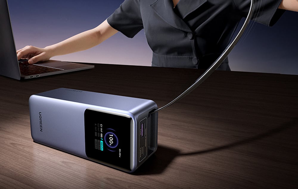 Powerbank leżący na ciemnym biurku obok osoby pracującej na laptopie, z wyciągniętym długim kablem ładującym.