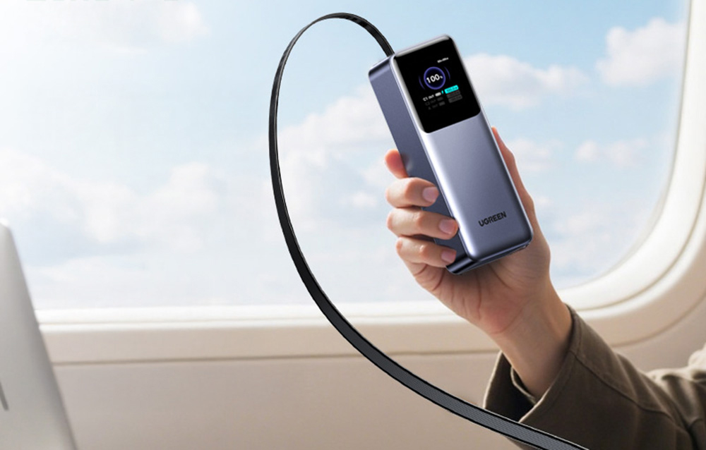 Dłoń trzymająca powerbank z aktywnym wyświetlaczem LCD na tle okna samolotu.