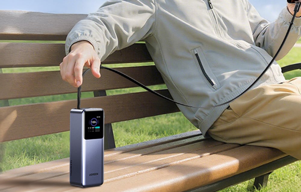 Powerbank stojący na ławce w parku, podłączony kablem do smartfona trzymanego przez siedzącą osobę.