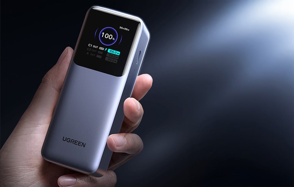 Zbliżenie na powerbank trzymany w dłoni, z wyraźnie widocznym stanem naładowania 100% na cyfrowym ekranie.