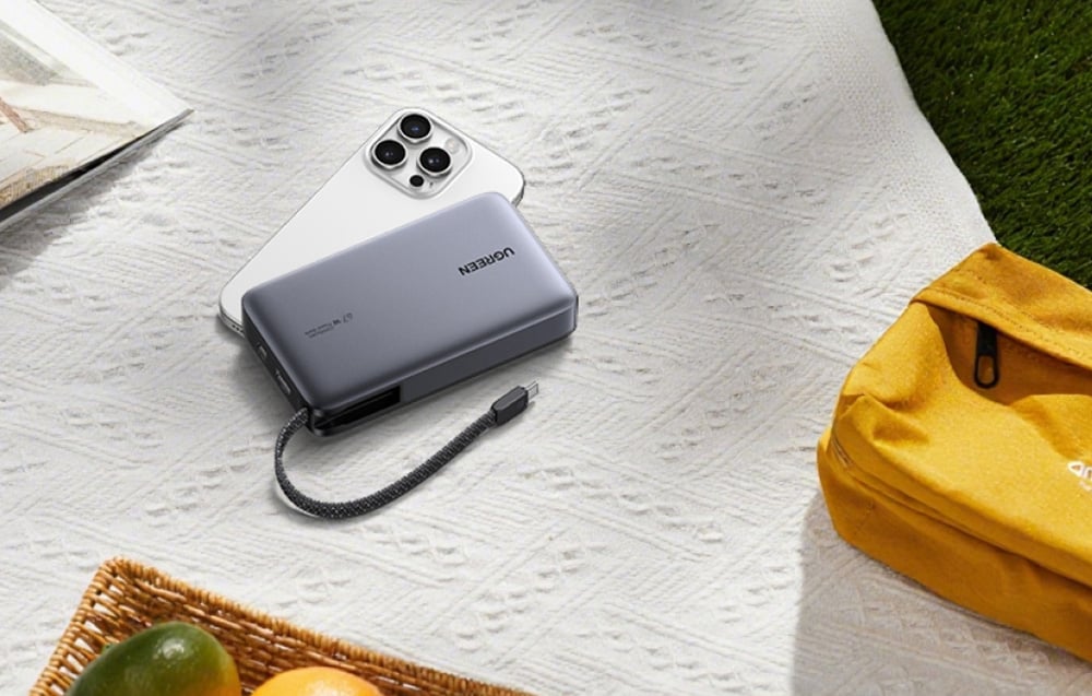 Powerbank Ugreen i iPhone leżące na kocu piknikowym obok żółtej torby, sugerujące wykorzystanie produktu w podróży i na zewnątrz.