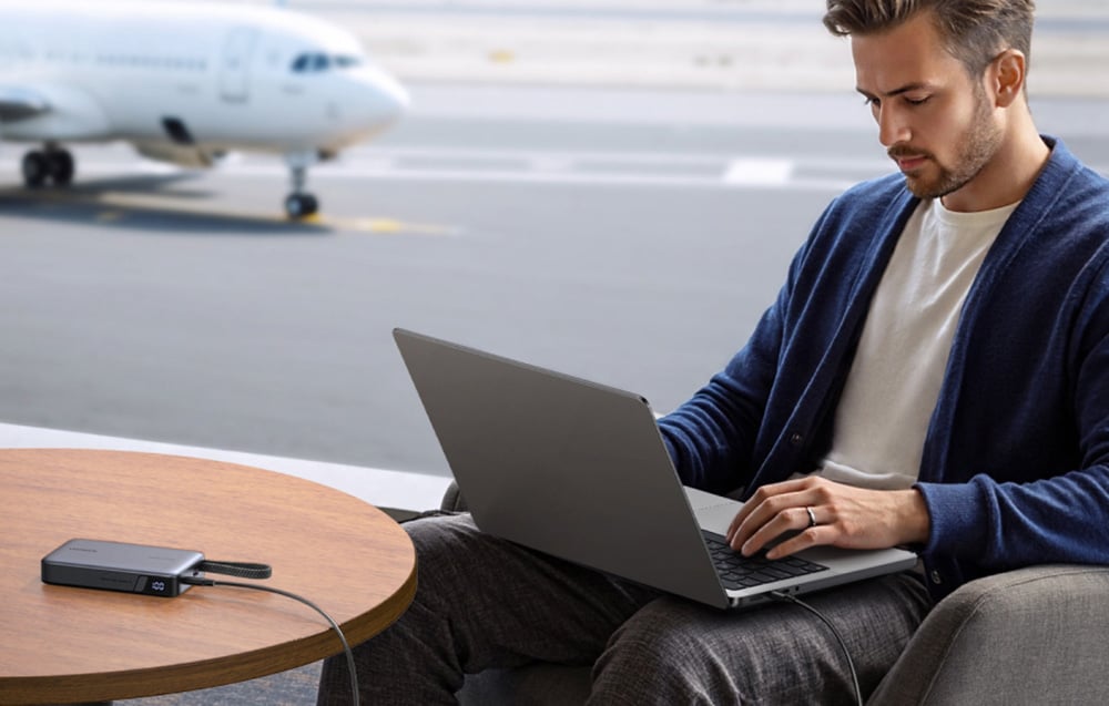 Mężczyzna pracujący na laptopie w terminalu lotniskowym, ładujący urządzenie z powerbanka Ugreen. W tle widoczny samolot.