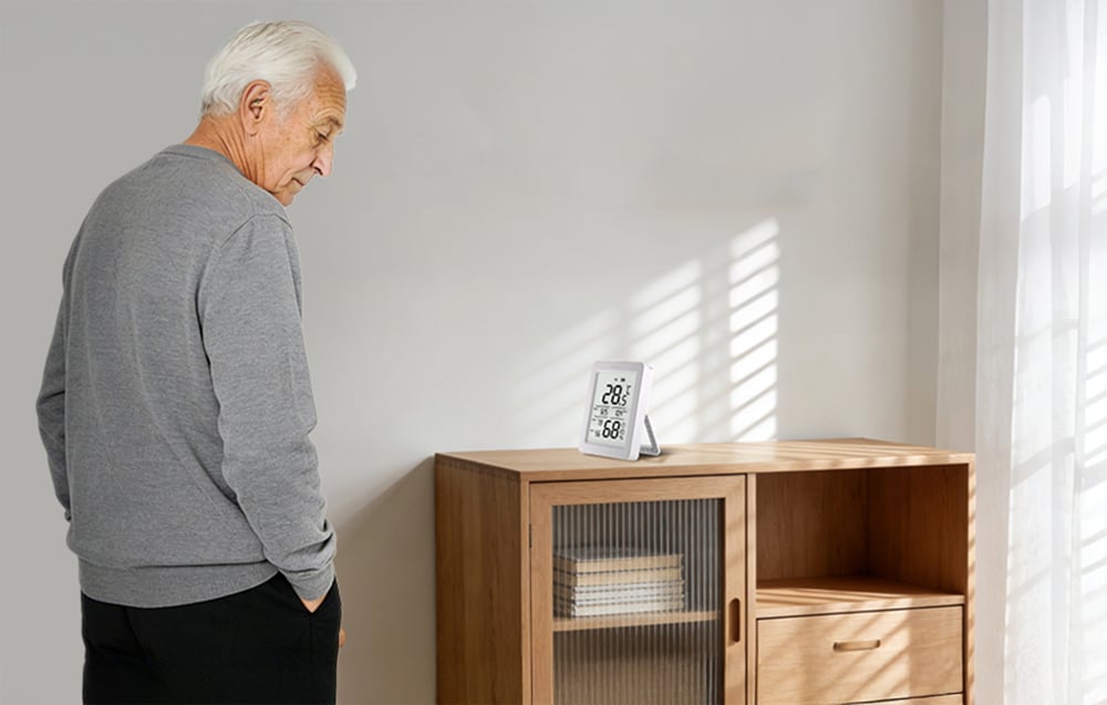 Starszy mężczyzna spogląda na czujnik temperatury i wilgotności ustawiony na komodzie w jasnym pokoju.