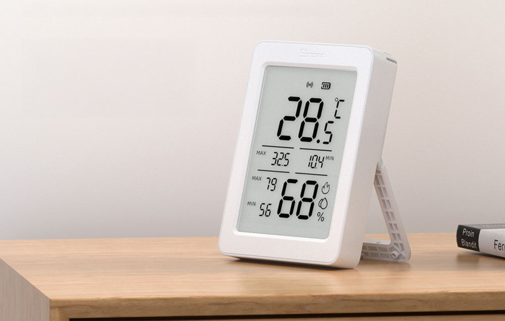 Czujnik Sonoff AirGuard TH stoi na drewnianej półce i pokazuje temperaturę oraz wilgotność na dużym ekranie LCD.
