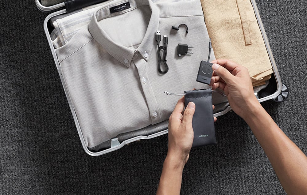 Otwarta walizka ze złożoną koszulą, ręce wkładające czarny adapter audio do woreczka, obok kabel USB i przejściówki