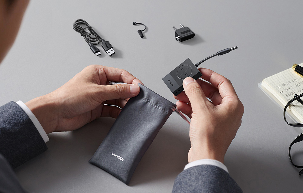 Ręce trzymają adapter audio i wkładają go do czarnego woreczka, obok kabel USB, przejściówka i notes