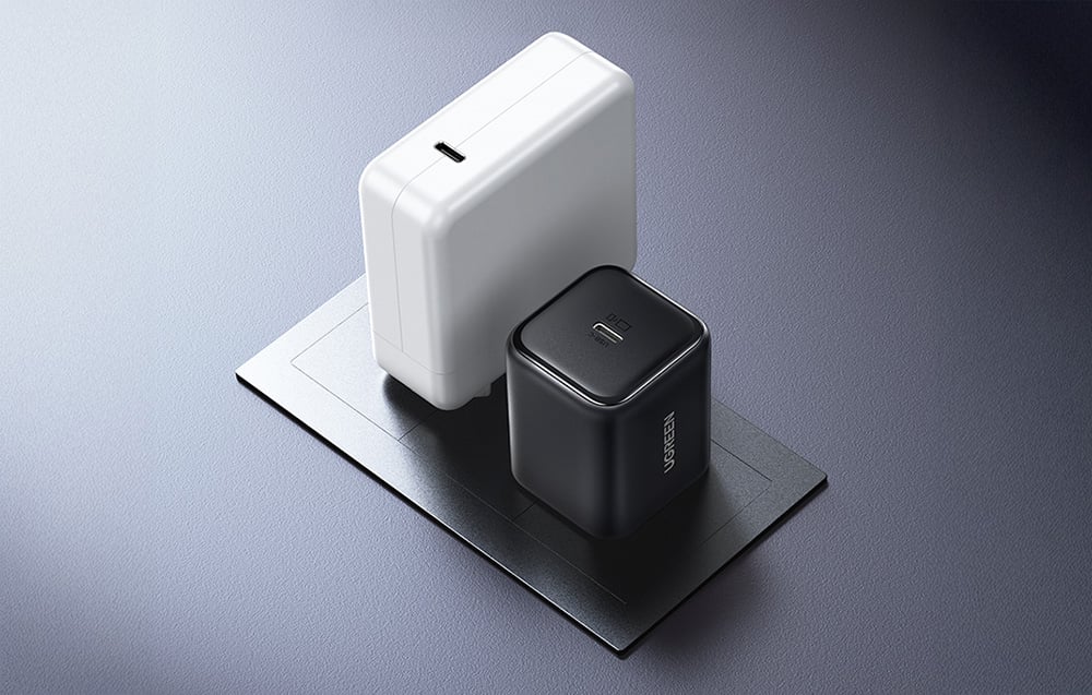 Czarna ładowarka UGREEN ustawiona obok większej białej ładowarki dla porównania rozmiaru na szarej powierzchni.