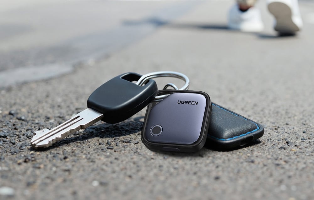 Lokalizator UGREEN przypięty do breloka z kluczami i etui leży na asfaltowej drodze.