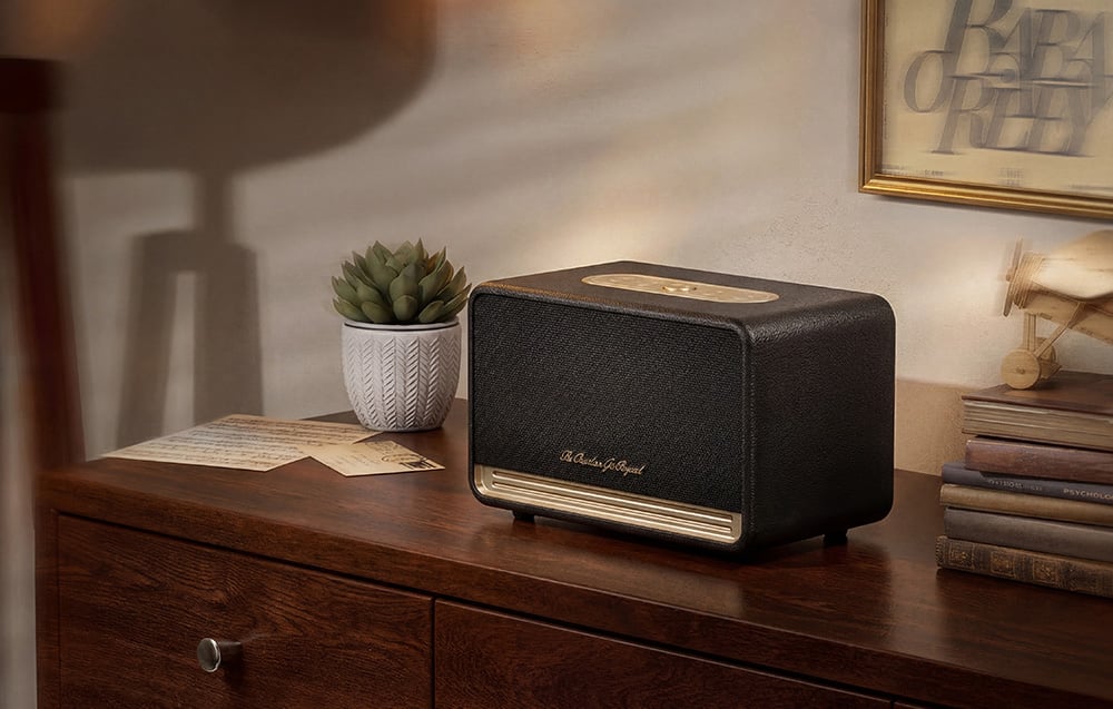 Czarny głośnik ze złotymi akcentami stoi na drewnianej komodzie obok książek i rośliny w doniczce