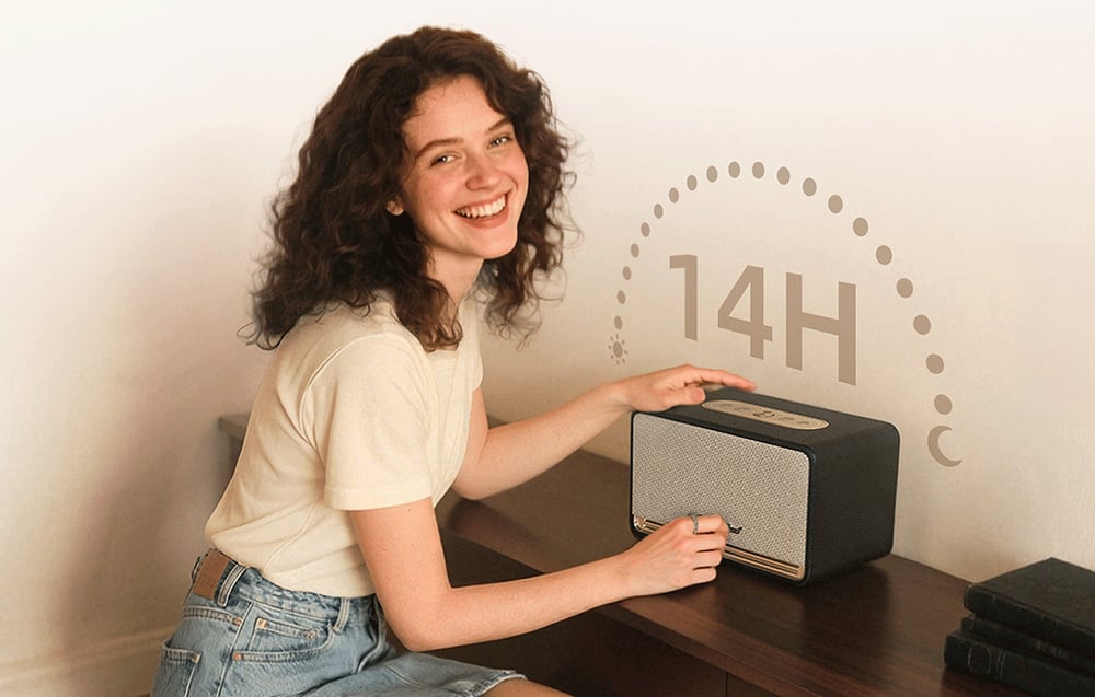 Uśmiechnięta kobieta korzysta z głośnika QCY leżącego na drewnianej komodzie, nad głośnikiem napis 14H