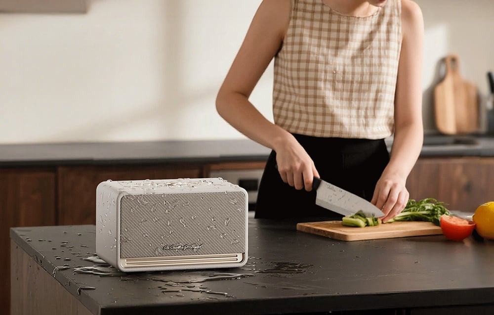 Kobieta kroi warzywa przy wyspie kuchennej, po lewej leży biały głośnik z kroplami wody