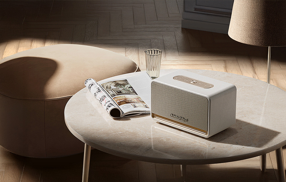 Biały głośnik ze złotymi akcentami leży w salonie na jasnym stoliku obok gazety i szklanki