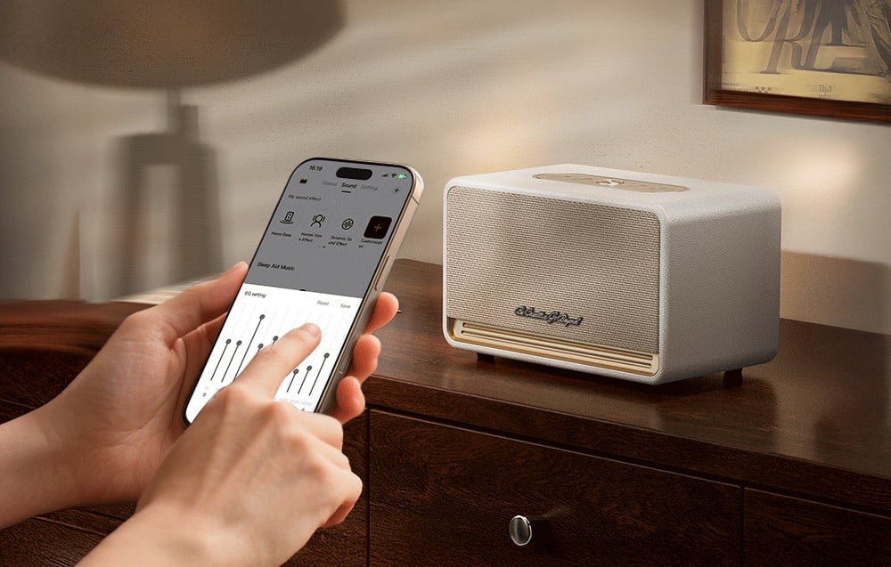 Trzymany w dłoni smartfon z włączoną aplikacją do sterowania dźwiękiem, po prawej biały głośnik na drewnianej komodzie