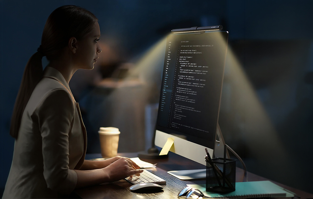 Woman sitting at a desk typing on a keyboard looking at a monitor with a lamp attached