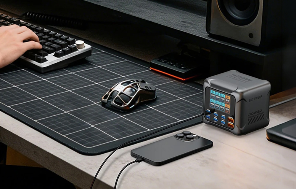 Încărcător pe un birou cu un smartphone conectat lângă un mouse de computer, cu o mână pe tastatură în stânga