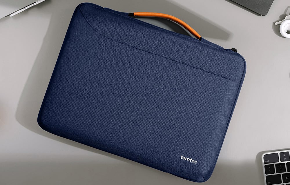 Granatowe etui tomtoc z pomarańczowym uchwytem, leżące płasko na jasnoszarym biurku obok klawiatury