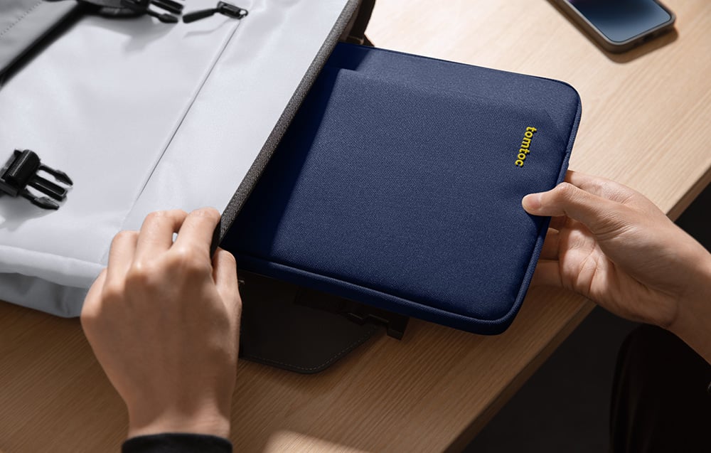 Ręce wkładające granatowe etui tomtoc do otwartego szarego plecaka na drewnianym biurku