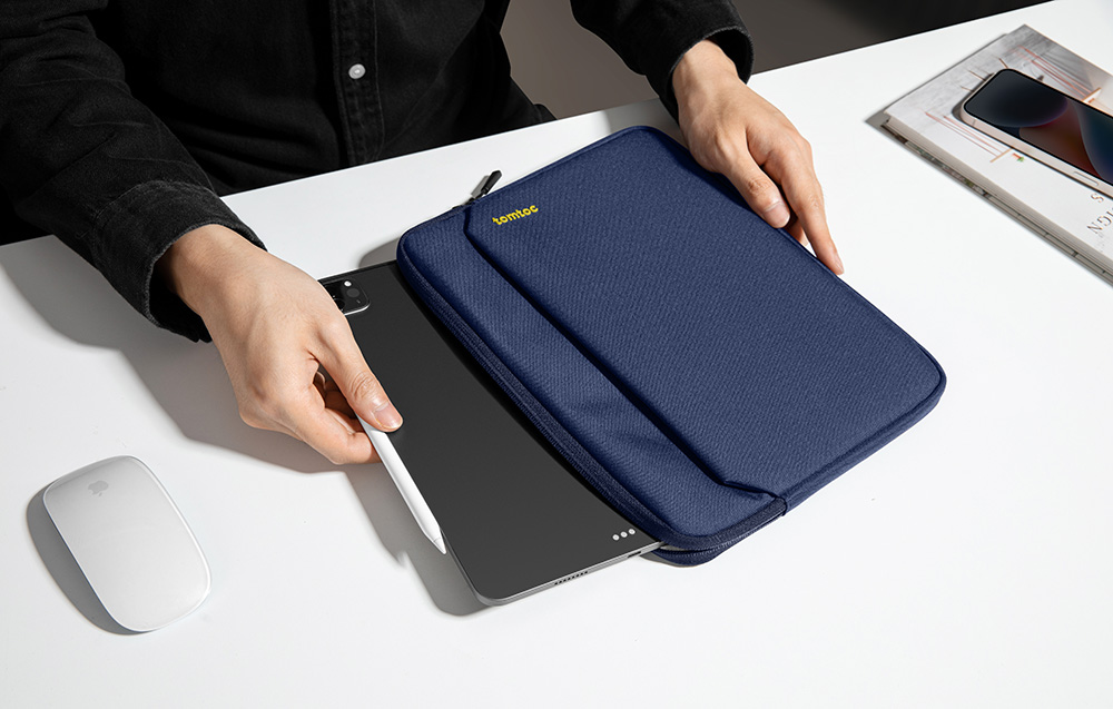 Ręce mężczyzny w czarnej koszuli wkładające iPada i Apple Pencil do granatowego etui tomtoc, na biurku mysz i telefon