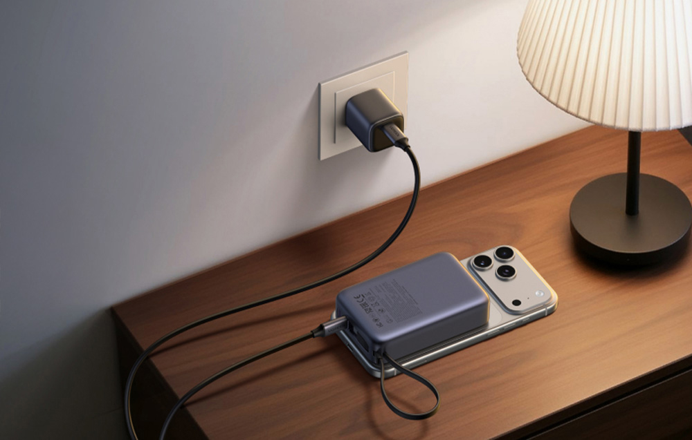 Szary powerbank magnetyczny Ugreen podłączony do wtyczki i ładujący bezprzewodowo smartfon leżący na szafce
