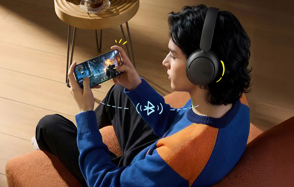 Mężczyzna w niebiesko-pomarańczowym swetrze siedzi na fotelu i gra w grę na smartfonie, mając założone czarne słuchawki