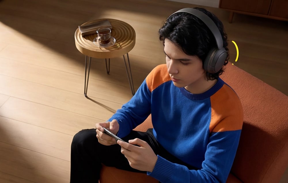 Mężczyzna w niebiesko-pomarańczowym swetrze siedzi na fotelu i trzyma smartfon w dłoniach, mając założone słuchawki