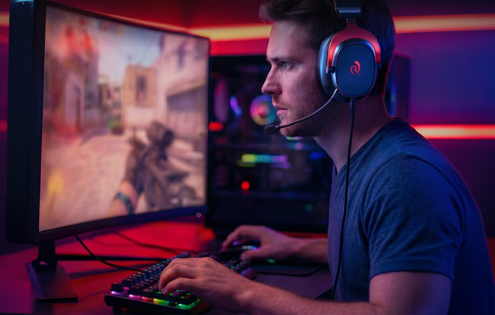 Mężczyzna w słuchawkach gamingowych grający w strzelankę FPS na zakrzywionym monitorze w pokoju z czerwonym i niebieskim oświetleniem