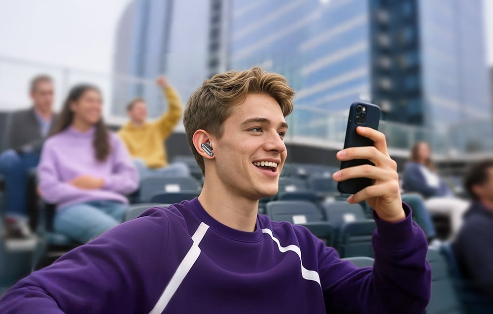 Młody mężczyzna w fioletowej bluzie siedzący na trybunach, trzymający smartfon i korzystający ze słuchawki