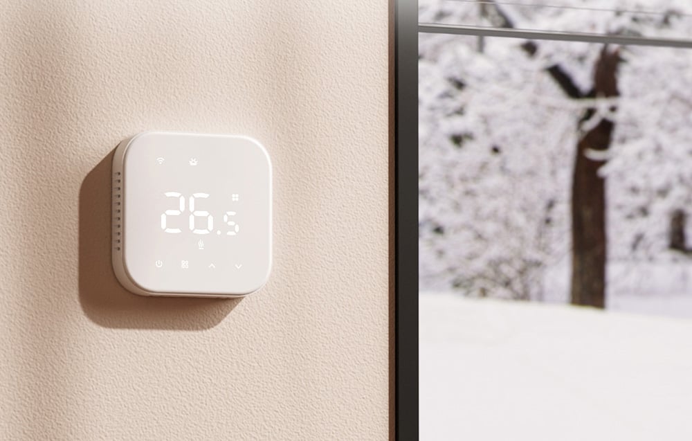 Biały inteligentny termostat Meross na beżowej ścianie obok okna z zimowym krajobrazem