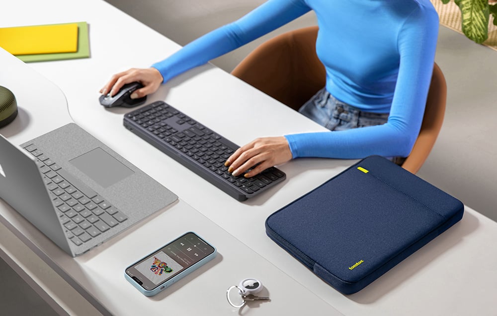Woman working at a desk with a laptop, keyboard, and navy Tomtoc sleeve