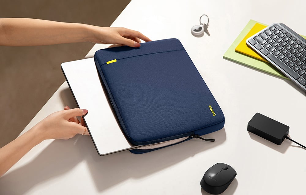 Hands placing a silver laptop into a navy Tomtoc sleeve on a white desk surface