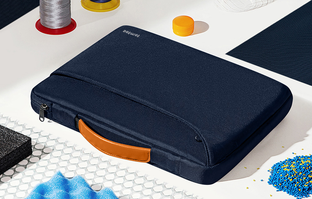 Granatowa torba na laptopa z pomarańczowym uchwytem leżąca na stole obok akcesoriów krawieckich i granulatu