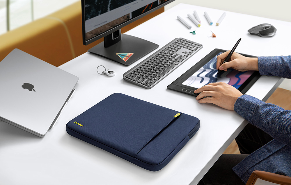Ciemnoniebieskie etui na laptopa tomtoc na białym biurku z monitorem, klawiaturą, tabletem graficznym i laptopem Apple