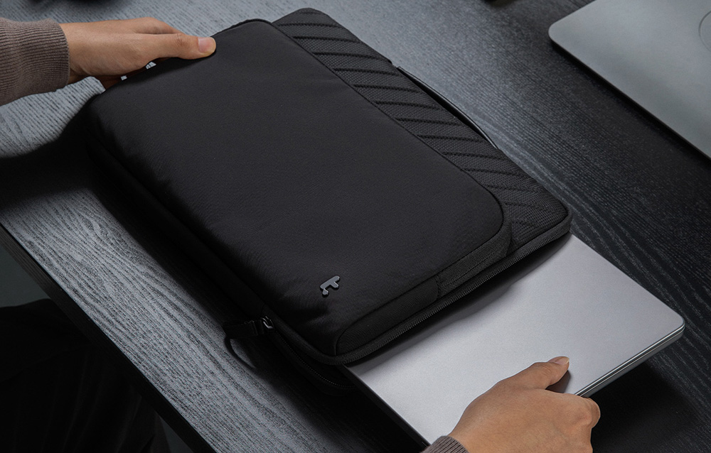 Ręce wysuwają srebrnego laptopa z czarnej torby z ukośnymi prążkami na ciemnym biurku