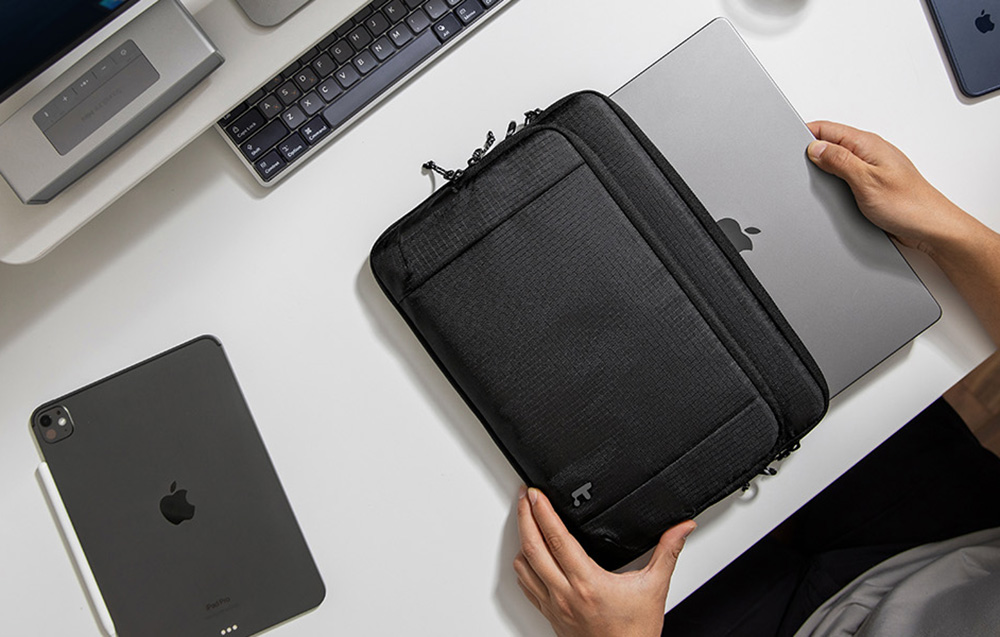 Ręce wyjmują srebrny laptop z czarnej torby na białym biurku z tabletem i klawiaturą