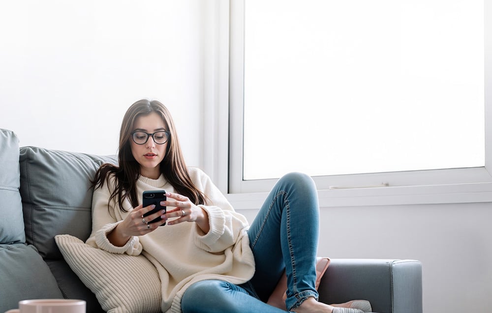 Žena v brýlích sedí na pohovce se smartphonem v ruce