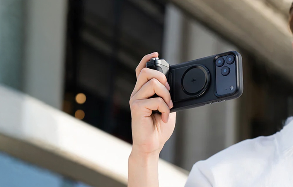 A hand holding a smartphone with a black grip while taking a photo outdoors