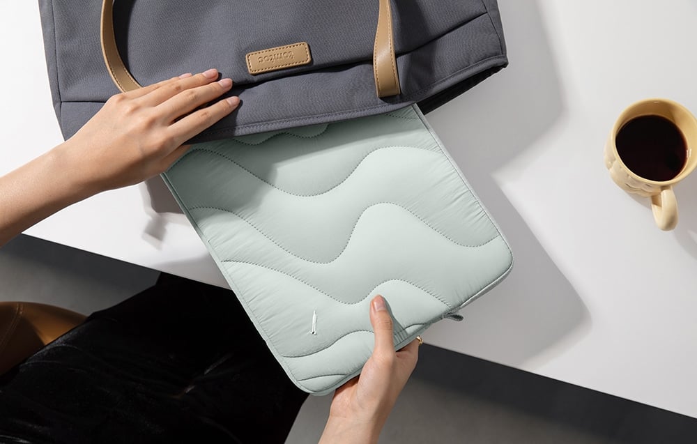 O persoană pune o husă quilted mint într-o geantă gri cu logo tomtoc, stând pe un blat alb lângă o cafea