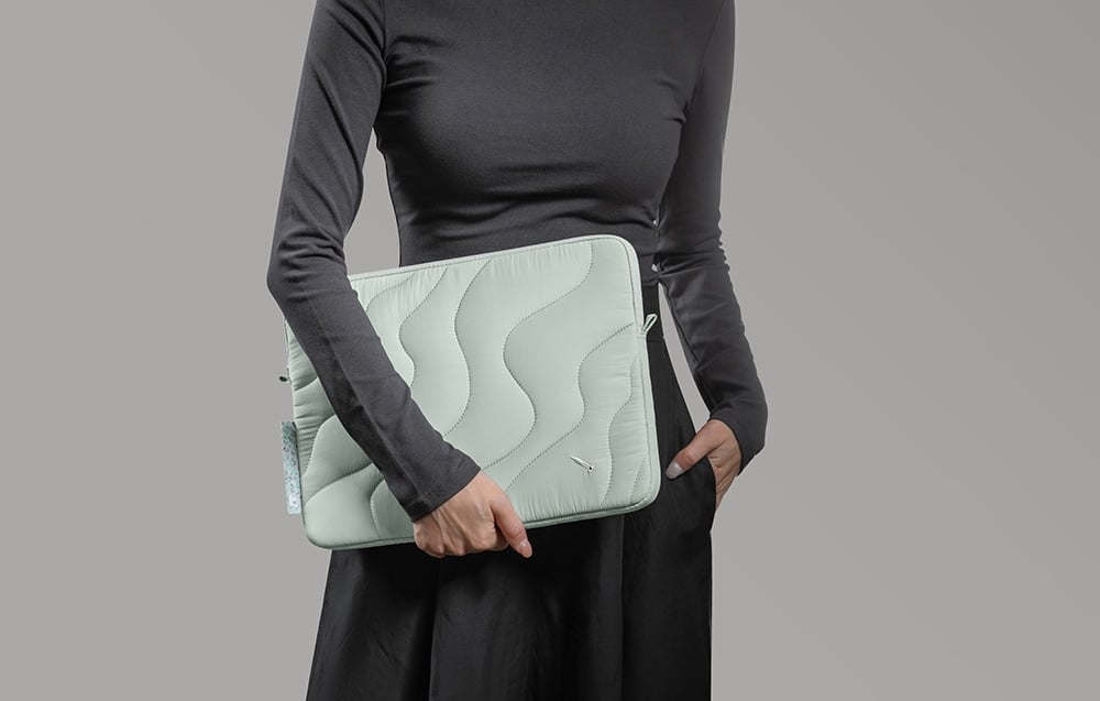 O femeie îmbrăcată în negru ține în mâini o husă quilted mint pentru laptop pe un fundal gri