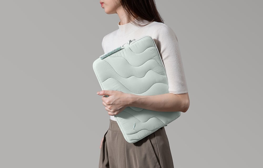 O femeie îmbrăcată în pulover alb ține sub braț o husă quilted mint pentru laptop pe un fundal deschis