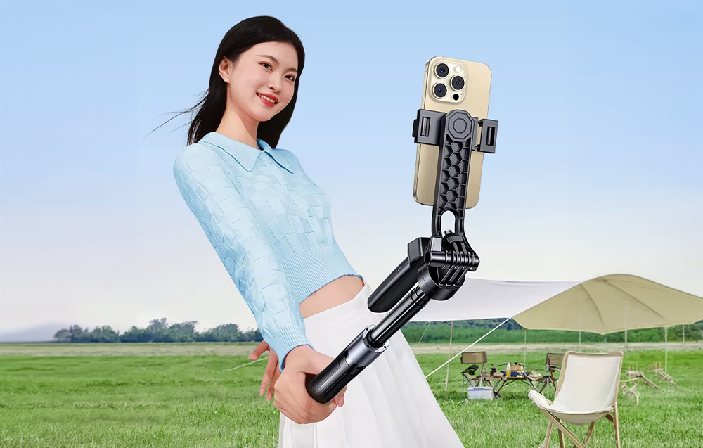 Smiling woman holding a selfie stick with a smartphone against the backdrop of a clearing and a tent