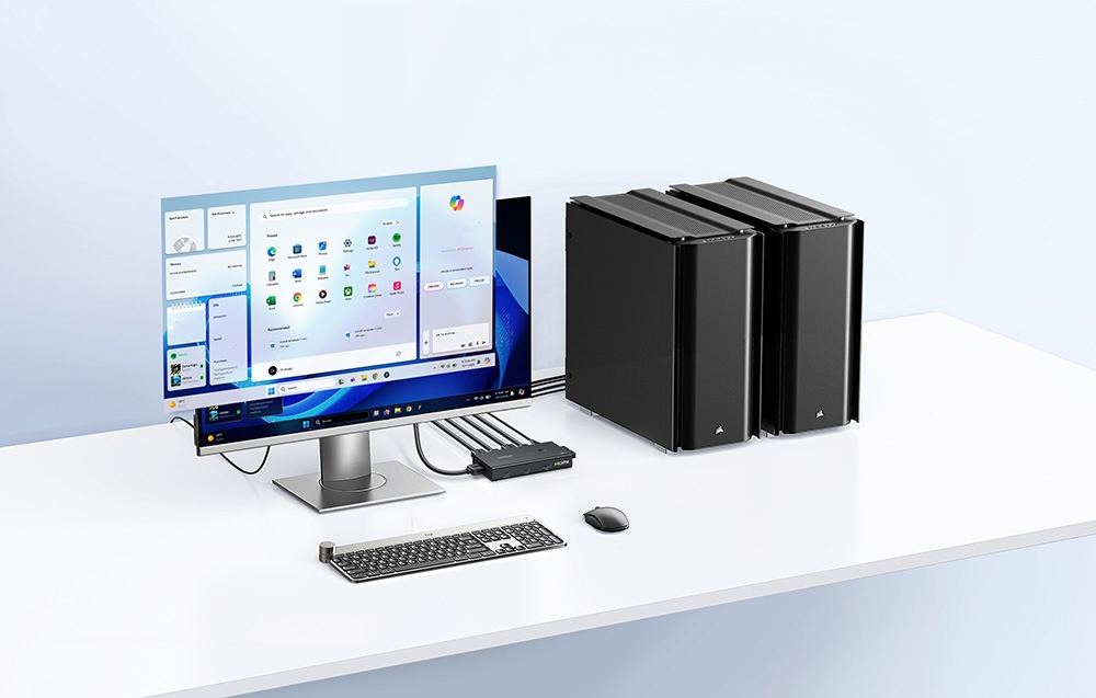 Pe un birou alb se află un monitor computer și două carcase de computer negre, așezate una lângă alta. În spatele monitorului se află un mic comutator cu cabluri conectate