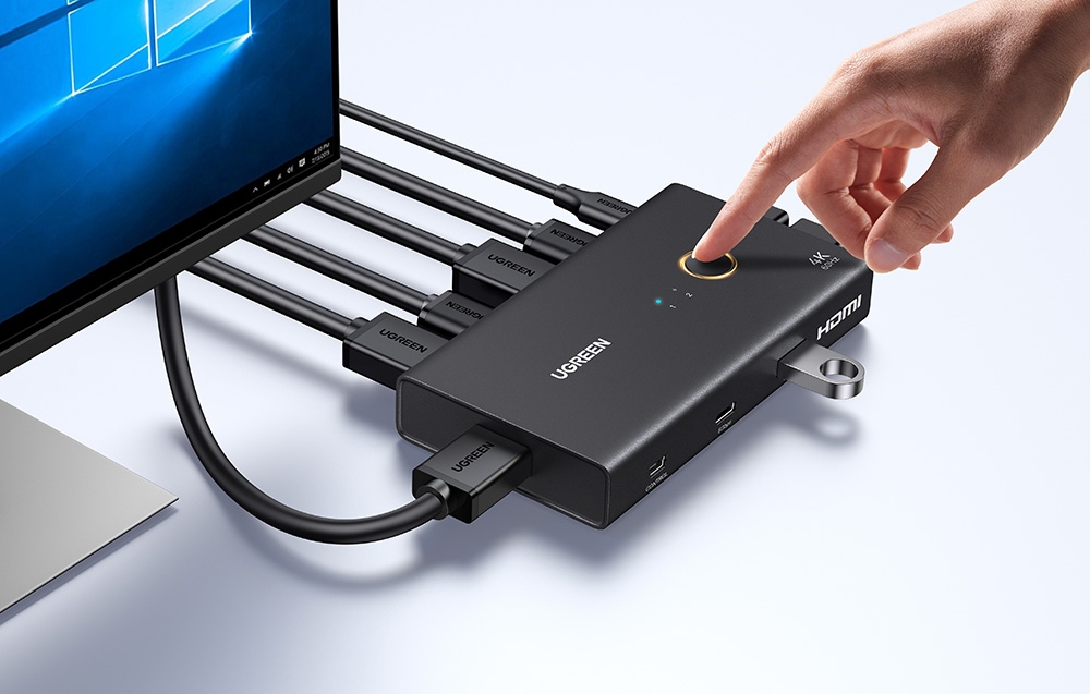 Un deget apasă un buton rotund pe carcasa superioară a comutatorului Ugreen. Pe lateral și pe spate sunt conectate mai multe cabluri negre și un stick USB