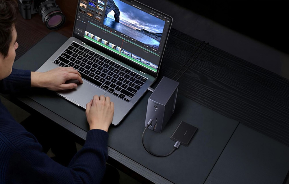 Widok z góry na mężczyznę pracującego na laptopie z uruchomionym programem do montażu wideo. Obok komputera leży mała stacja dokująca, do której podłączony jest płaski dysk zewnętrzny
