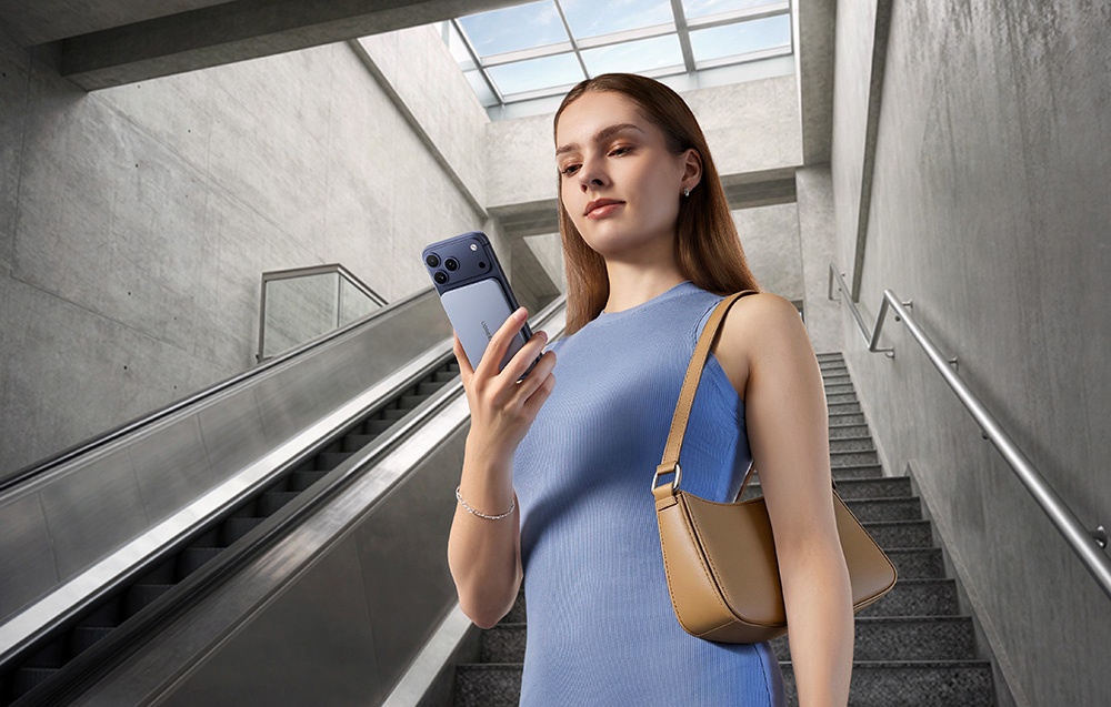 A woman in a light blue dress stands on concrete stairs next to an escalator holding a smartphone with a power bank attached to its case