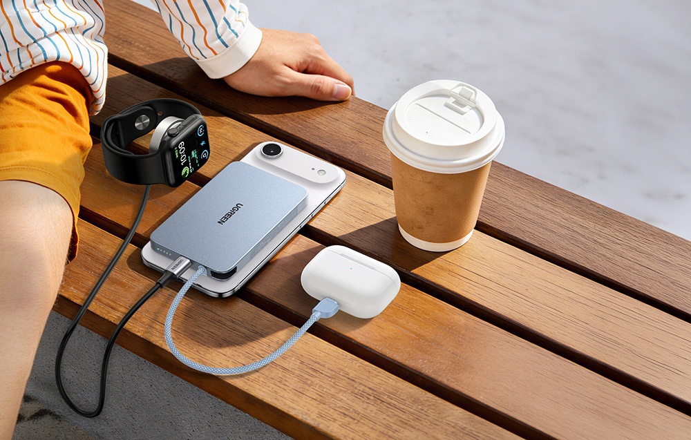 Smartphone, TWS headphones, and smartwatch connected to the Ugreen power bank lying on a bench next to a coffee cup, with a person sitting on the left