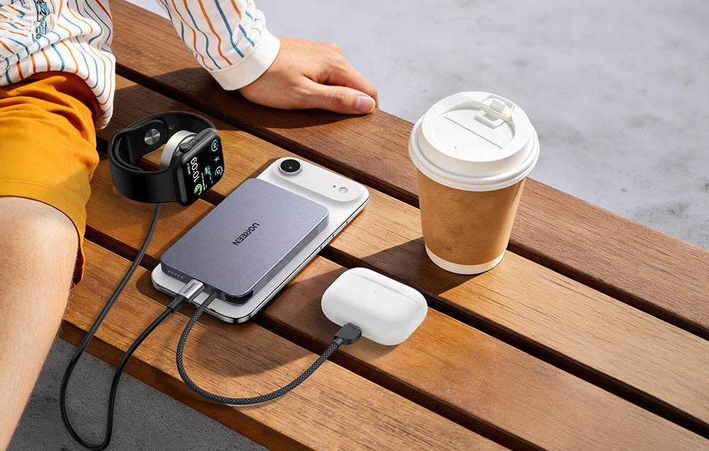 Smartphone, TWS headphones, and smartwatch connected to the Ugreen powerbank lying on a bench next to a coffee cup, with a person sitting on the left