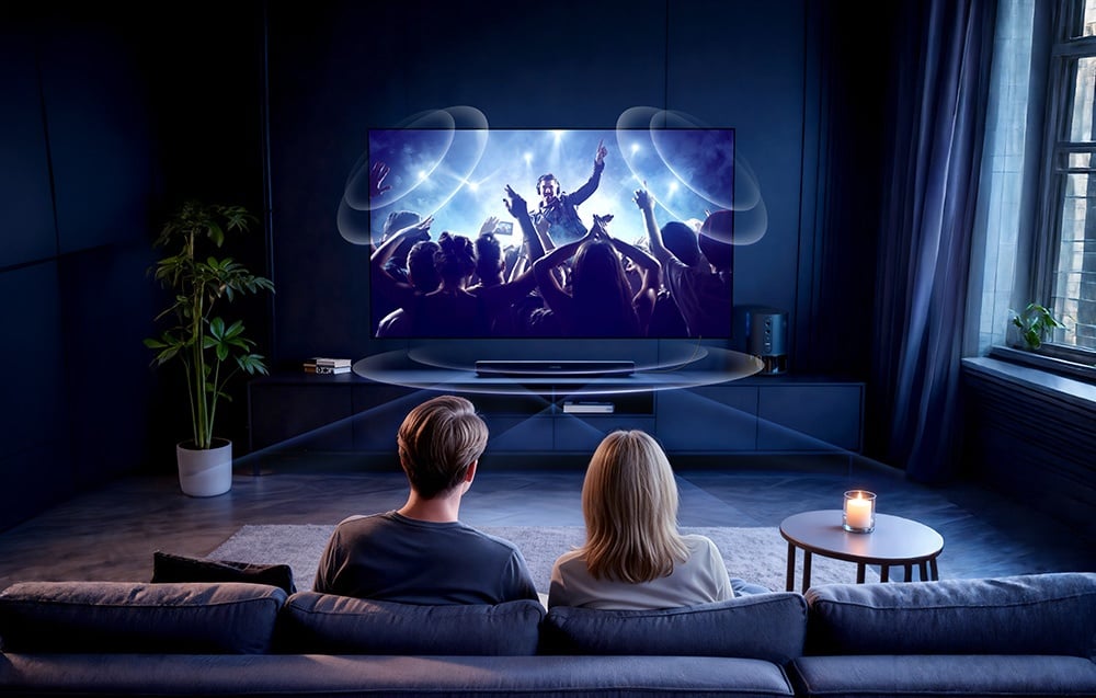 Dwie osoby siedzą tyłem do kadru na dużej kanapie przed telewizorem wyświetlającym koncert muzyczny.